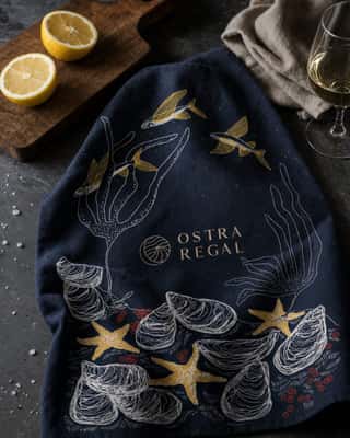Torchon bleu nuit collab Morgan Chouin 2025 étalé sur une ardoise, avec une planche en bois, deux demi-citrons et un verre de vin blanc