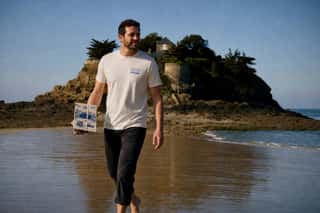 Homme en t-shirt Ostra Regal portant un coffret d'huîtres sur la plage, le Fort du Guesclin derrière lui